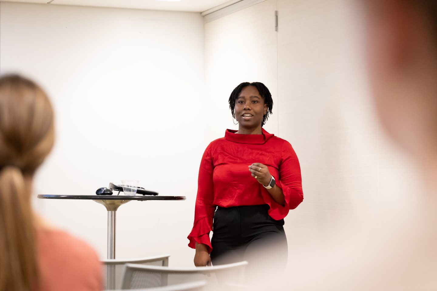 A Black woman student wearing a long-sleeved red blouse, rings, a wristwatch and black pants, is standing before an audience, making a presentation in a room with white walls. 
