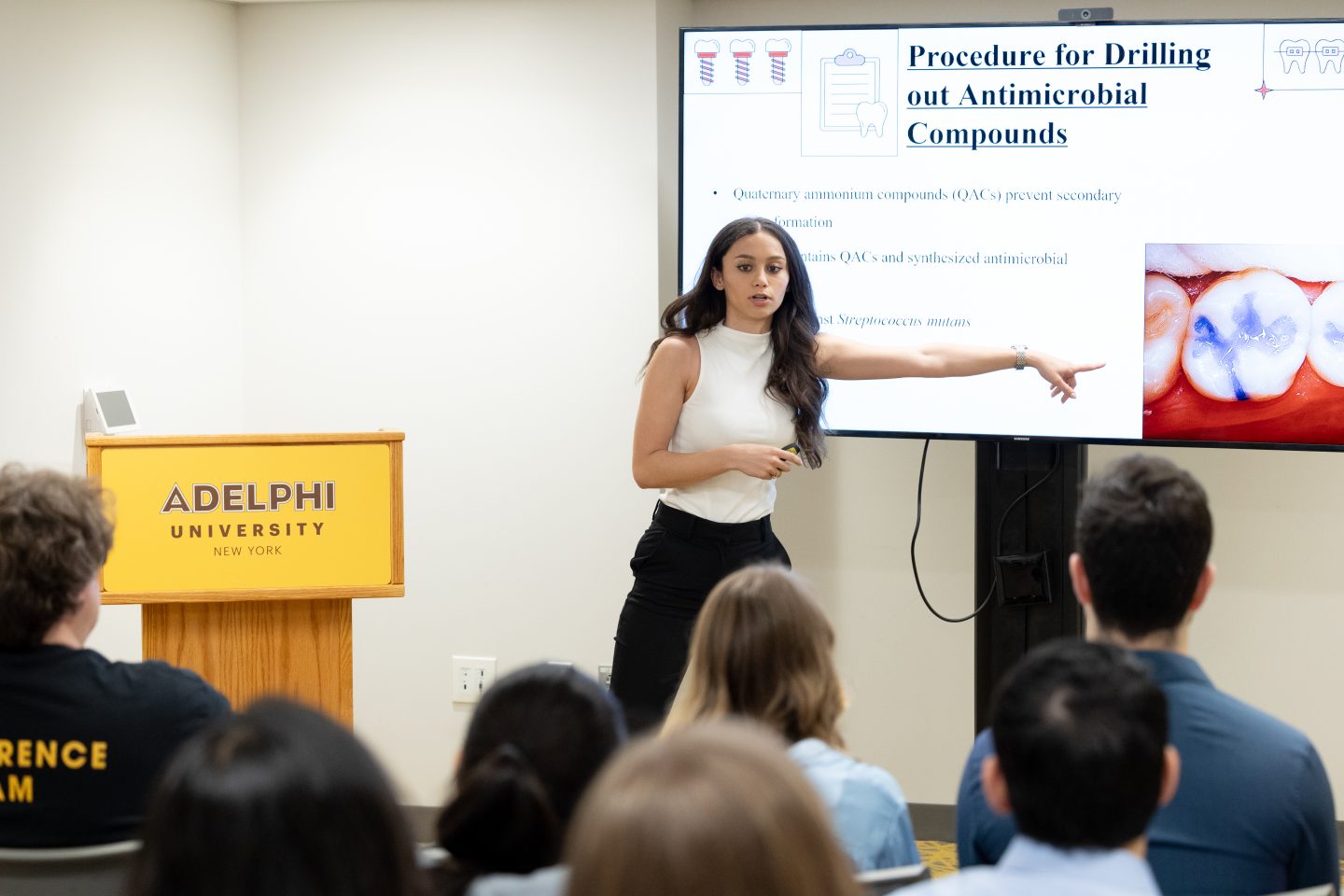 A female student with long dark brown hair, wearing a sleeveless white blouse and black slacks, is gesturing with her left arm toward a large video screen. To her right is a podium. In the foreground, we see the backs of seven audience members watching her presentation.