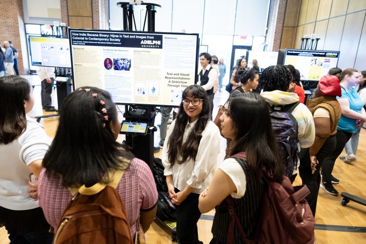 A female student with long dark brown hair, wearing a sleeveless white blouse and black slacks, is gesturing with her left arm toward a large video screen. To her right is a podium. In the foreground, we see the backs of seven audience members watching her presentation.