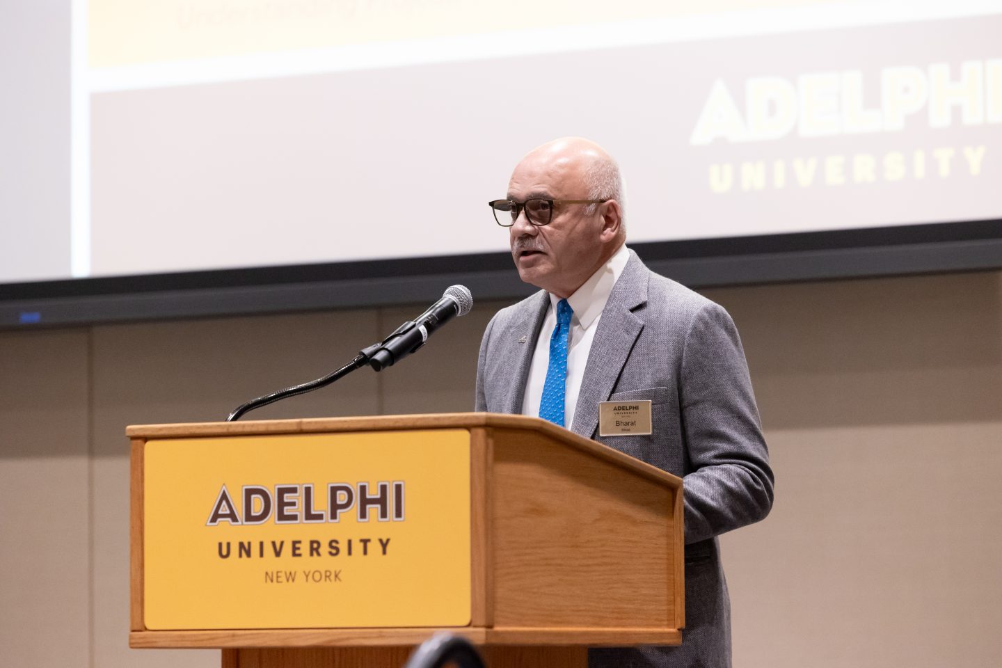 An adult man with short white hair, wearing a gray suit, a bright blue necktie and name tag, is standing at a podium and speaking into a microphone. A large video screen is in the background.