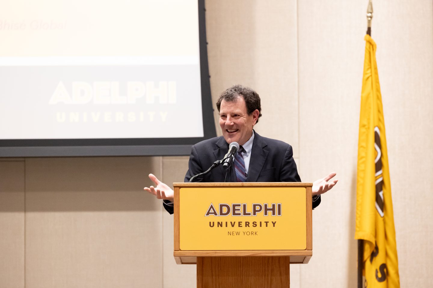 An adult man standing at a podium addressing the public. There is a yellow flag to his left and screen behind him.