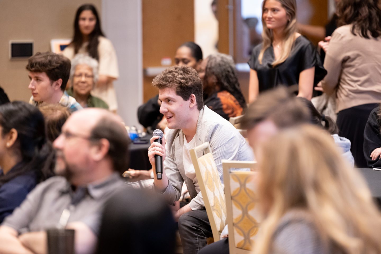 A male student with light brown hair is speaking into a handheld microphone in a room full of other event attendees—a mixture of young students and adult faculty. Most are seated, though some in the background are standing.