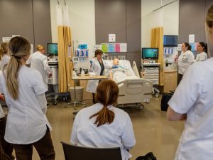 A group of nursing students wearing white uniforms attentively surrounding a patient mannequin in a simulated hospital room, providing care and support.