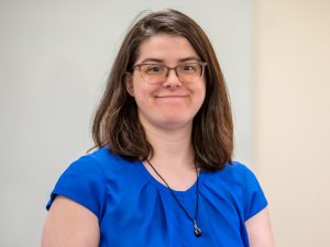 A woman with shoulder-length brown hair wearing glasses, a royal blue blouse, and a black necklace, smiling.