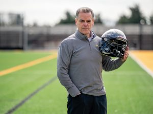 A man standing on a football field holding a football helmet in his left hand. He is wearing a grey quarter-zip pullover and dark pants. The field has green turf with yellow and black markings.