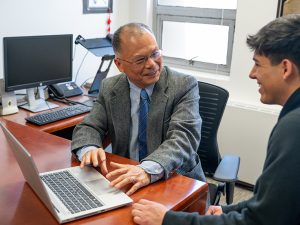 An older man in a gray blazer and blue tie sits at a desk with a laptop, smiling and talking with a younger man in a black hoodie in an office setting