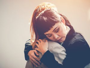 A close-up image of two people hugging each other in a solemn manner. One person has dark hair with bangs and a dark jacket and the other has long hair in a light-colored outfit