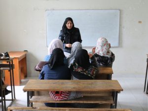 A female teacher in a black headscarf stands at the front of a classroom near a whiteboard, speaking to several younger students wearing headscarves who are seated at wooden desks