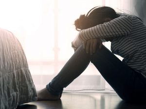 A female sitting on the floor of a bedroom with her knees pulled up to her chest, arms wrapped over her knees and head bowed down on her arms