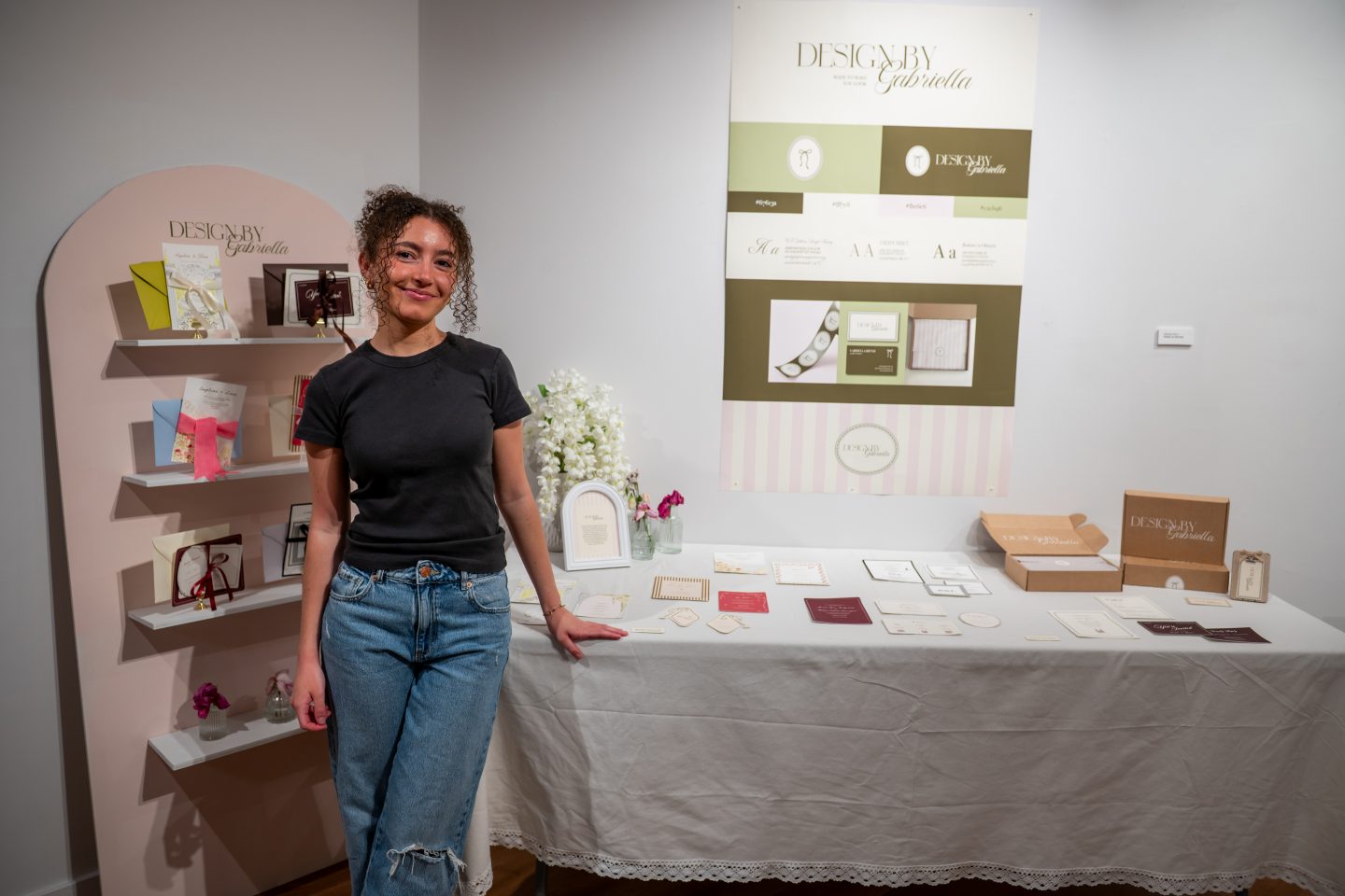 A woman in a black t-shirt and distressed jeans stands at a design display booth for "Design By Gabriella," showcasing a luxury branding and graphic design portfolio. Behind her is an arched rose-toned shelf unit featuring design samples, books, and decorative items. A large poster board displays the brand's logo, color palette, typography, and design applications. The white table in front displays printed branding materials including business cards, stationery, and packaging samples in neutral and earth tones.