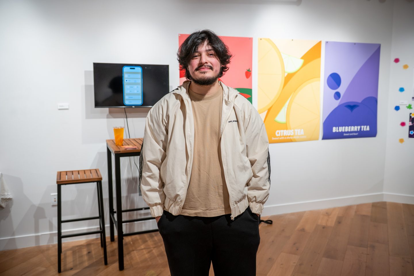 A man with dark hair and a mustache stands in front of a colorful research poster display featuring beverage designs labeled "Citrus Tea" and "Blueberry Tea" with vibrant illustrated graphics. Behind him is a small table with a mobile phone displaying an app interface mounted on a stand, and wooden bar stools. The display is set against a white gallery-style wall.
