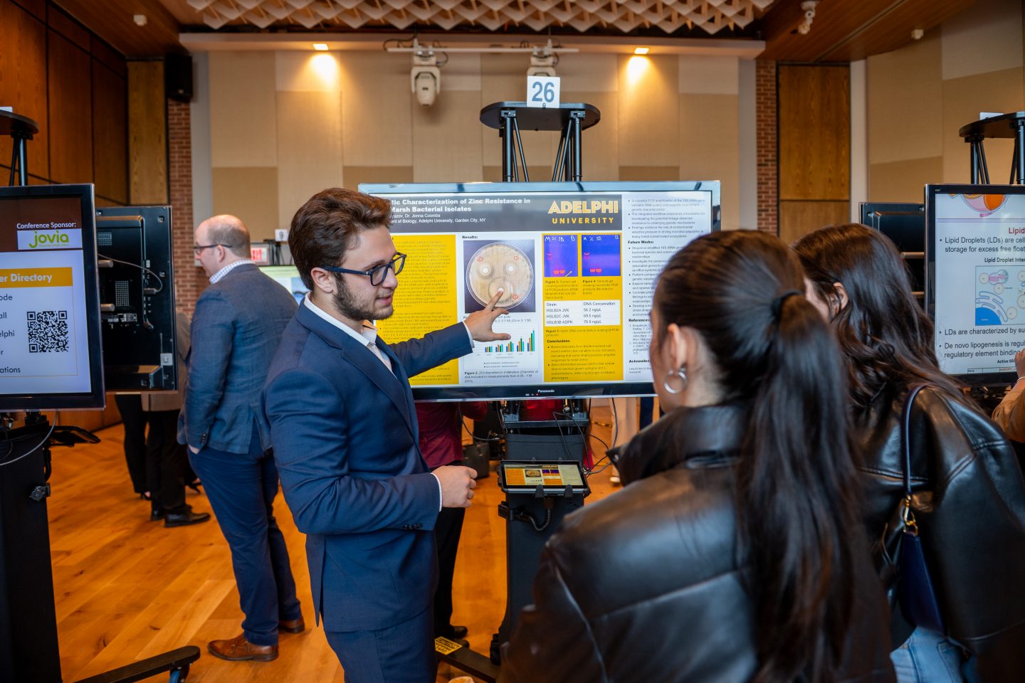 A male research student in a dark blue suit and glasses presents his Adelphi University research on "Molecular Characterization of Zinc Resistance in Gram-Negative Bacterial Isolates" to two visitors at a poster session. The poster displays yellow and blue sections with scientific imagery, charts, and data. A tablet device sits at the base of the display. Other researchers and attendees are visible in the background of the brick-walled venue with wooden ceiling details.