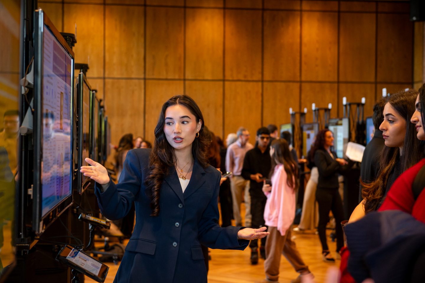 A young woman in a dark navy blazer presents her research at a poster session, gesturing toward a digital display screen mounted on a stand. She speaks to visitors gathered around her booth while other attendees view posters in the background of a wood-paneled venue.