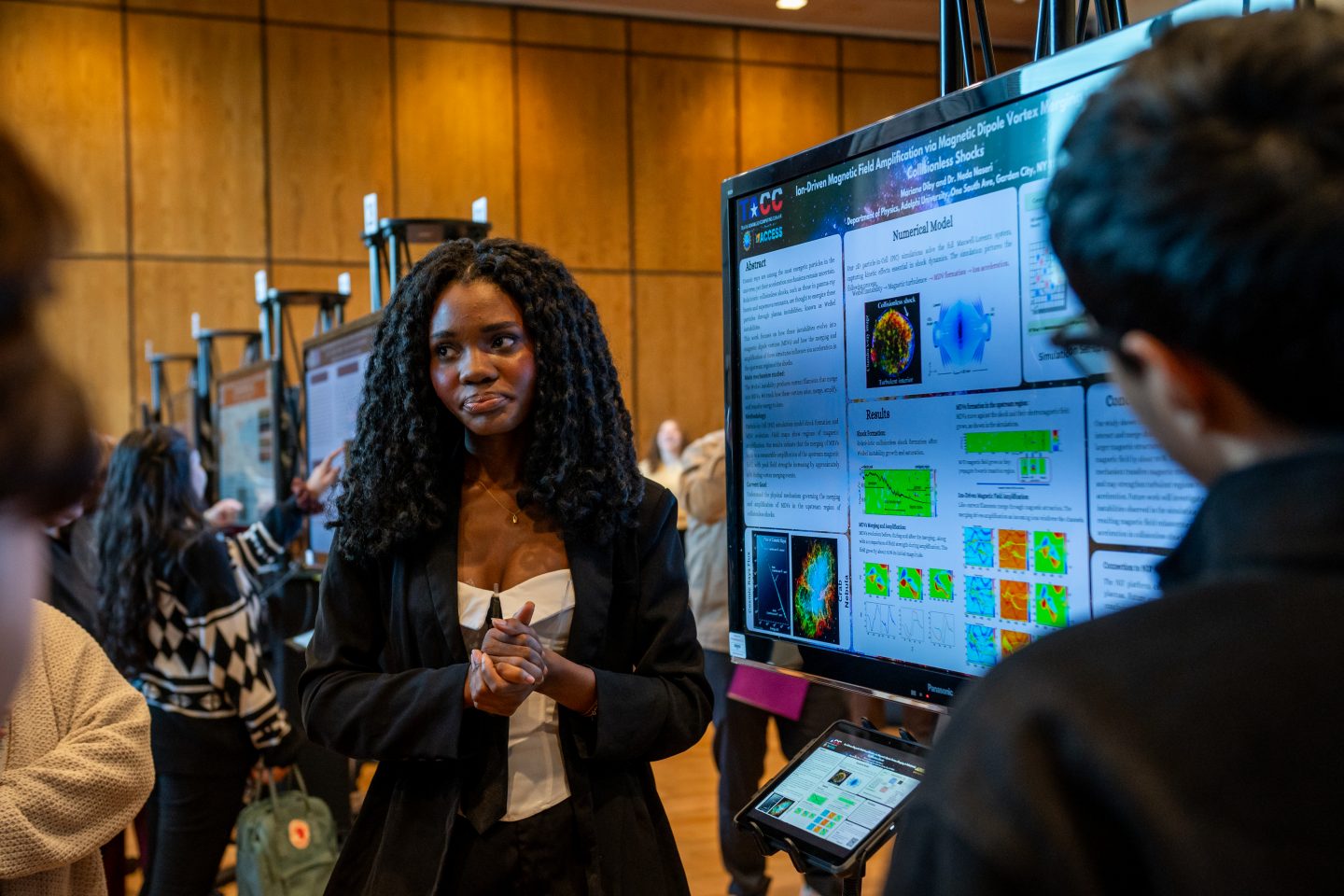 A female research student in a black blazer and cream top presents her work to a visitor at a research poster session. Her poster displays scientific research on magnetic field amplification with brain imaging visualizations and heat maps in various colors. A tablet device sits at the base of the poster. Multiple other research posters are visible in the background of the wood-paneled venue, with other attendees and researchers present.
