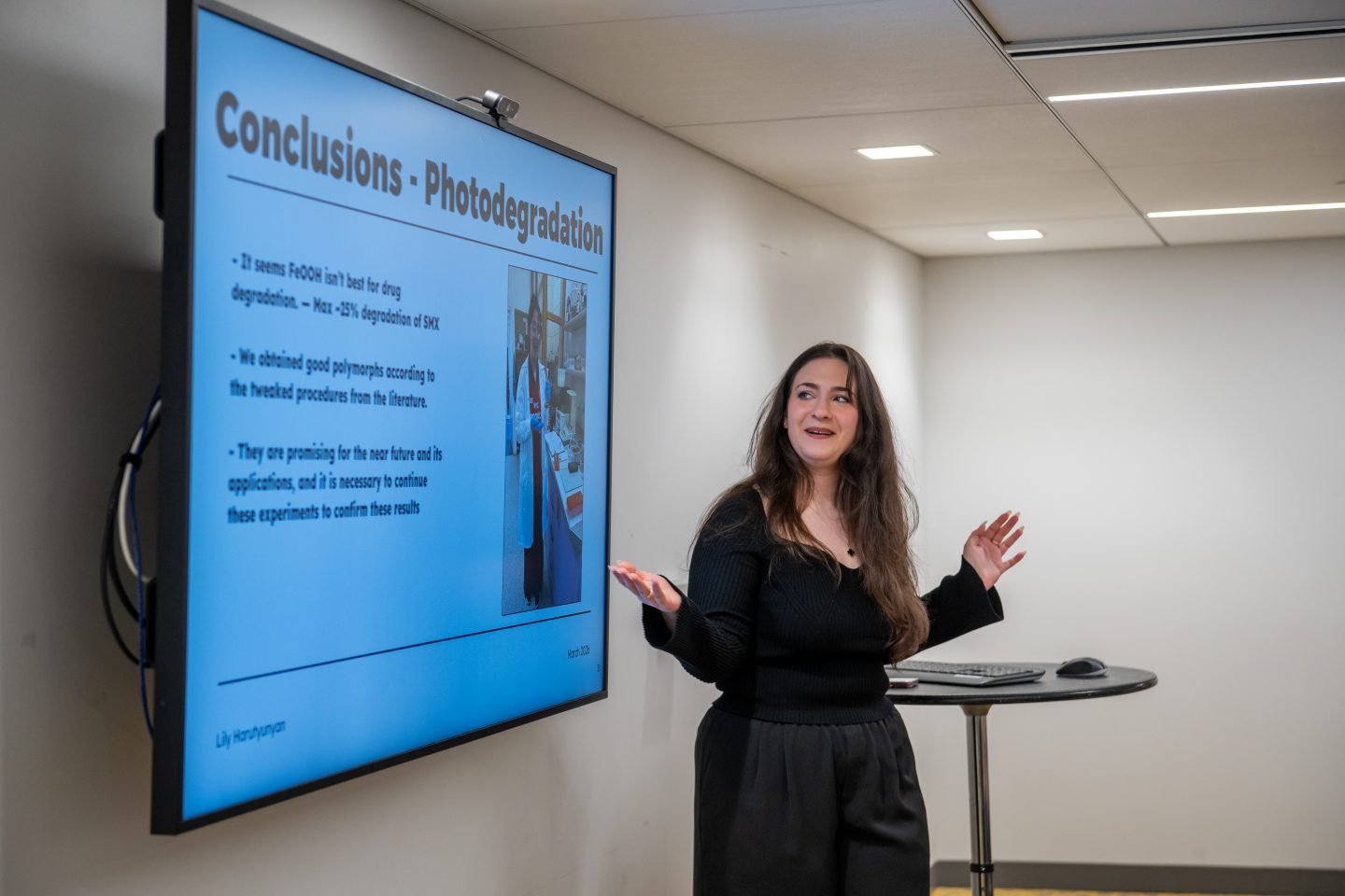 A woman in a black dress gestures while presenting research findings on a large blue screen display. The screen shows a "Conclusions • Photodegradation" slide with text discussing drug degradation results and laboratory equipment imagery. A small table with a computer mouse and keyboard is visible beside her in a modern presentation room with recessed ceiling lights.