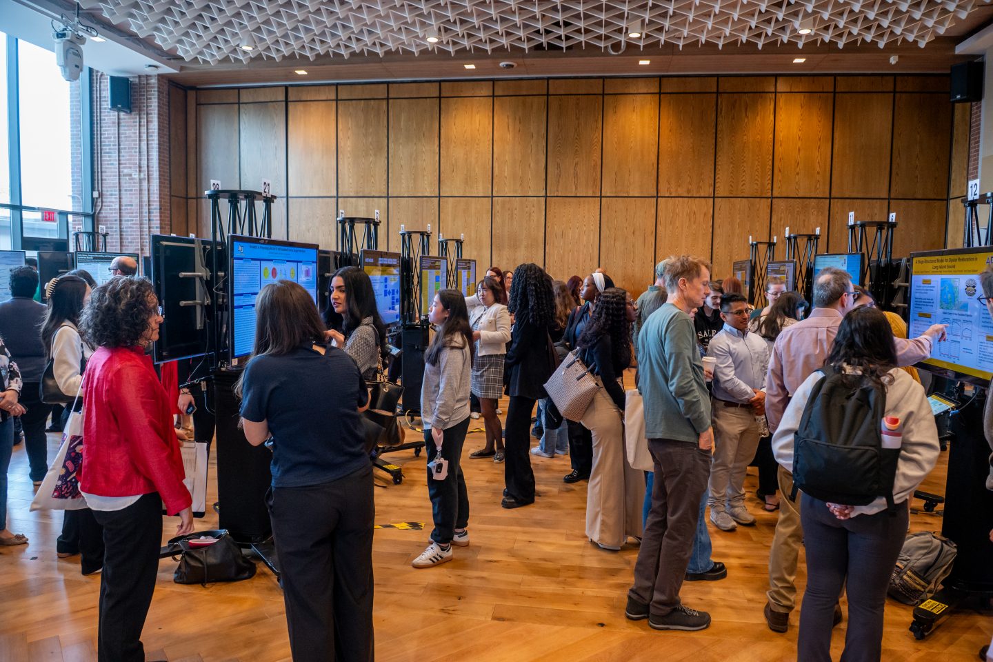 A large crowd of attendees mingles throughout a research symposium hall, viewing multiple research posters displayed on stands with blue screens. The modern venue features a distinctive geometric ceiling, wood paneling, and wooden flooring. Visitors in casual to business casual attire engage with researchers at various poster stations.