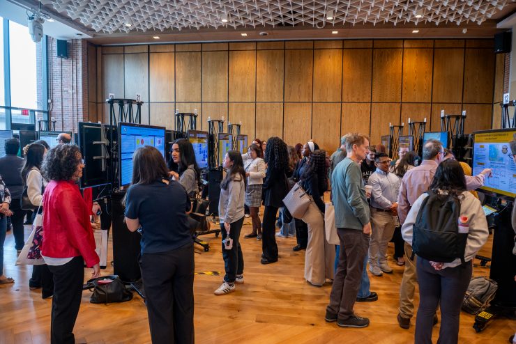 A male research student in a dark blue suit and glasses presents his Adelphi University research on "Molecular Characterization of Zinc Resistance in Gram-Negative Bacterial Isolates" to two visitors at a poster session. The poster displays yellow and blue sections with scientific imagery, charts, and data. A tablet device sits at the base of the display. Other researchers and attendees are visible in the background of the brick-walled venue with wooden ceiling details.