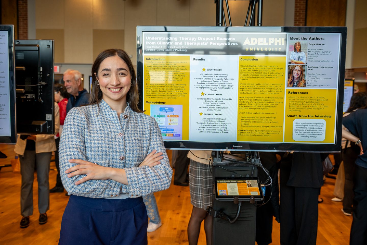 A smiling graduate student in a blue patterned shirt stands with arms crossed in front of a research poster at Adelphi University. The poster displays her research on "Understanding Therapy Dropout Reasons from Clients' and Therapists' Perspectives" with sections for Introduction, Methodology, Results, and Conclusion. A "Meet the Authors" panel appears on the right side of the poster. Other attendees are visible in the background of what appears to be a research day event in a brick-lined venue.