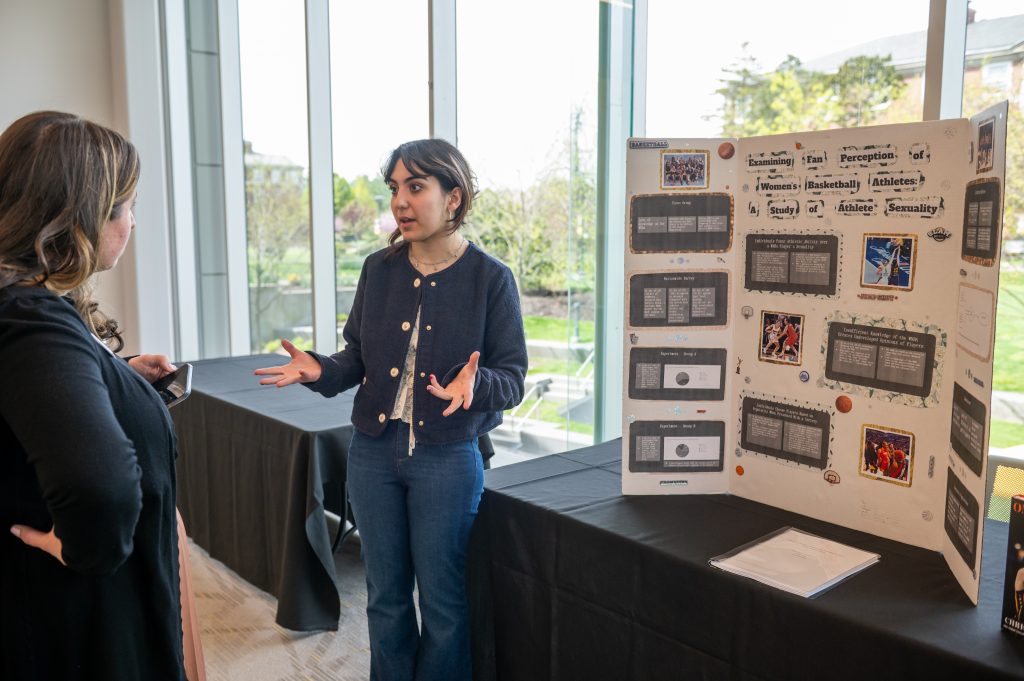 A student presenter explains her research poster about women's basketball and athlete perception to a visitor at the Scholarship & Creative Works Conference, with large windows overlooking campus grounds in the background.