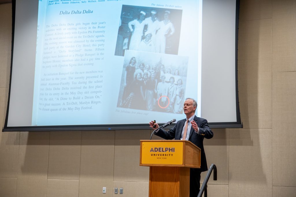 Nobel laureate Dr. William Kaelin speaks at an Adelphi University podium during a keynote presentation, gesturing while discussing a slide about Delta Delta Delta sorority history projected behind him.