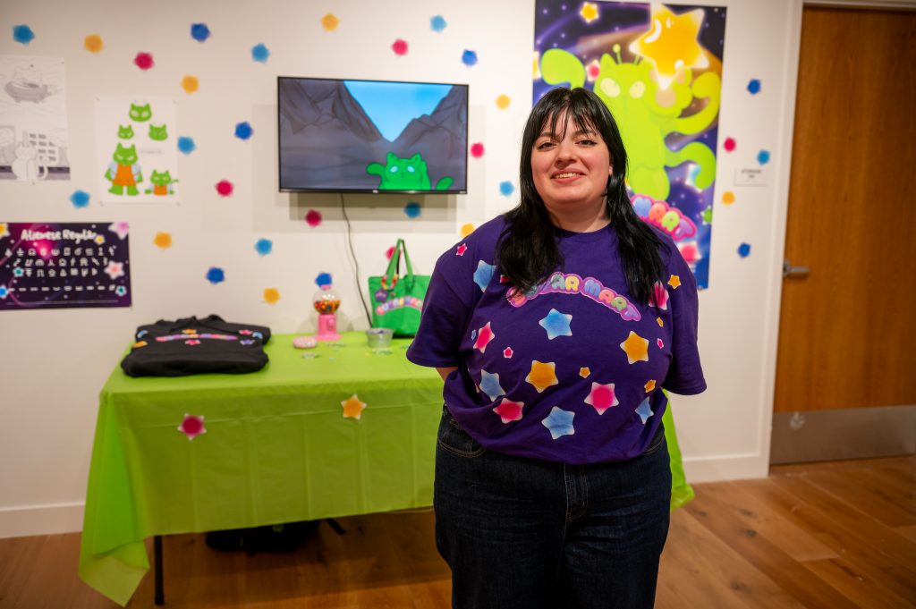 A graphic design student stands smiling in front of her colorful exhibition display featuring bright green and purple artwork, animated characters on a monitor, and playful decorative elements including colorful paper shapes hanging from the ceiling.