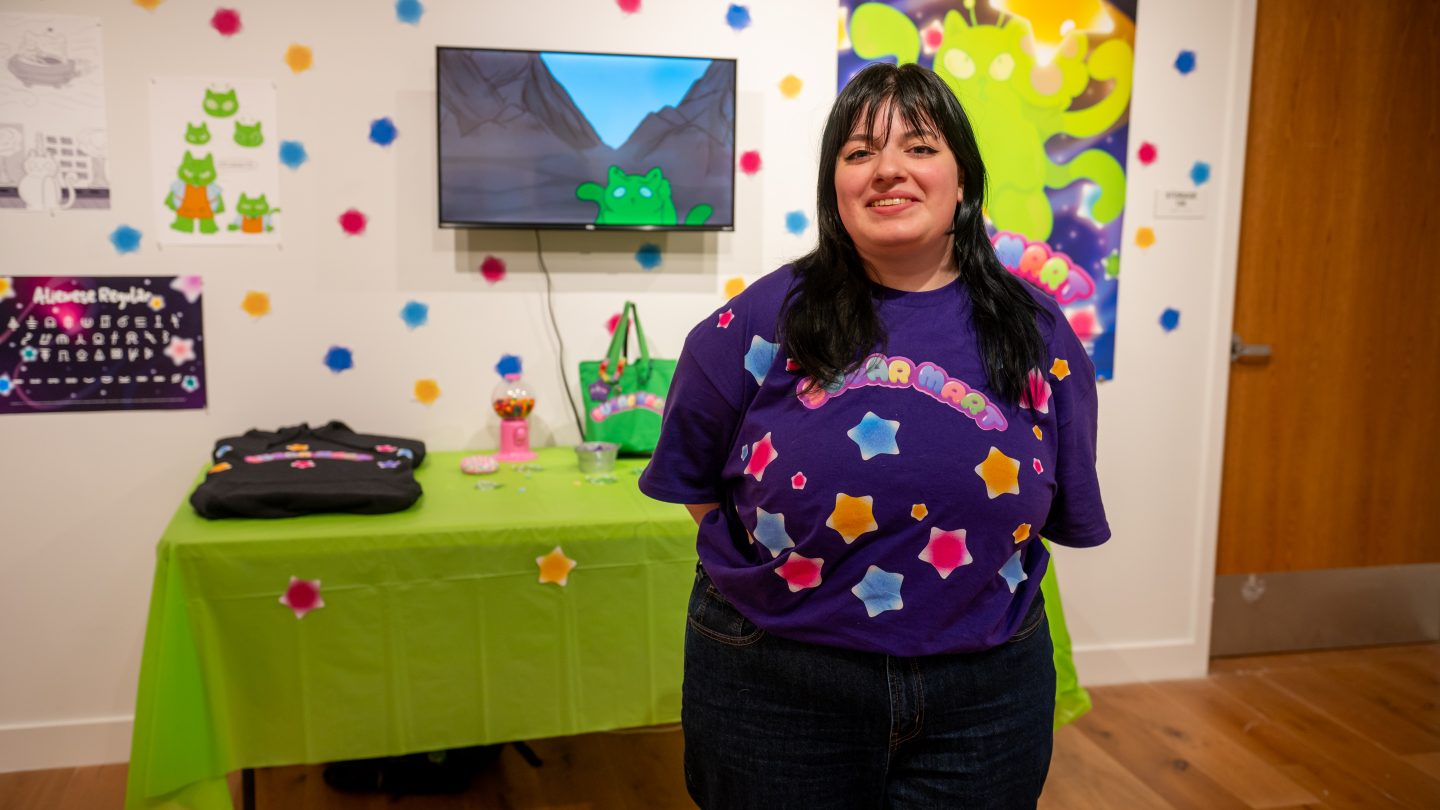 A graphic design student stands smiling in front of her colorful exhibition display featuring bright green and purple artwork, animated characters on a monitor, and playful decorative elements including colorful paper shapes hanging from the ceiling.