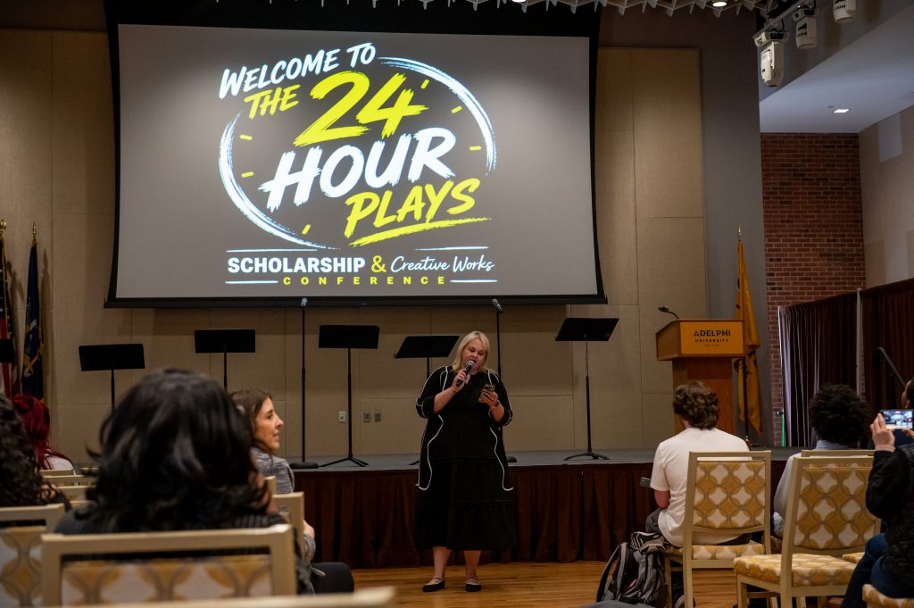 A speaker presents at the Scholarship & Creative Works Conference, standing at a podium in front of a large screen displaying "Welcome to the 24 Hour Plays: Scholarship & Creative Works Conference" with an Adelphi University podium visible.