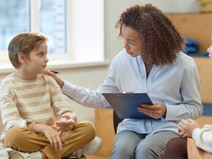 A counselor holding a clipboard talks to a young boy while sitting with a small group of children in a classroom setting.