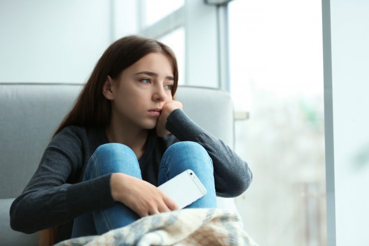 Upset teenage girl with smartphone sitting at window indoors.