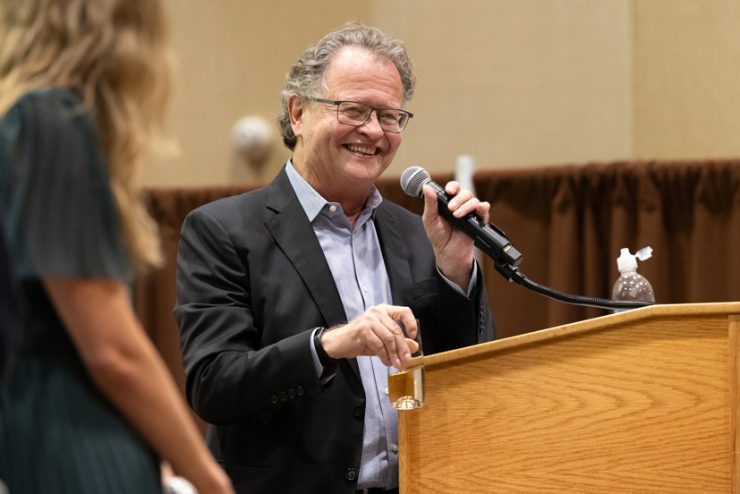 A man, smiling, holds a microphone at a lectern. A woman with her back to the camera stands on the left.