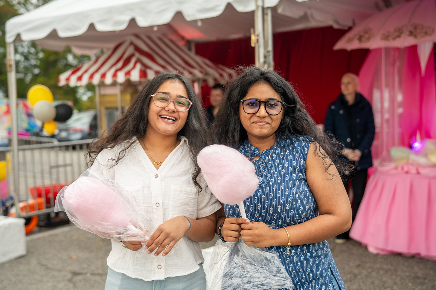 Two women share cotton candy, with a colorful carnival behind them.