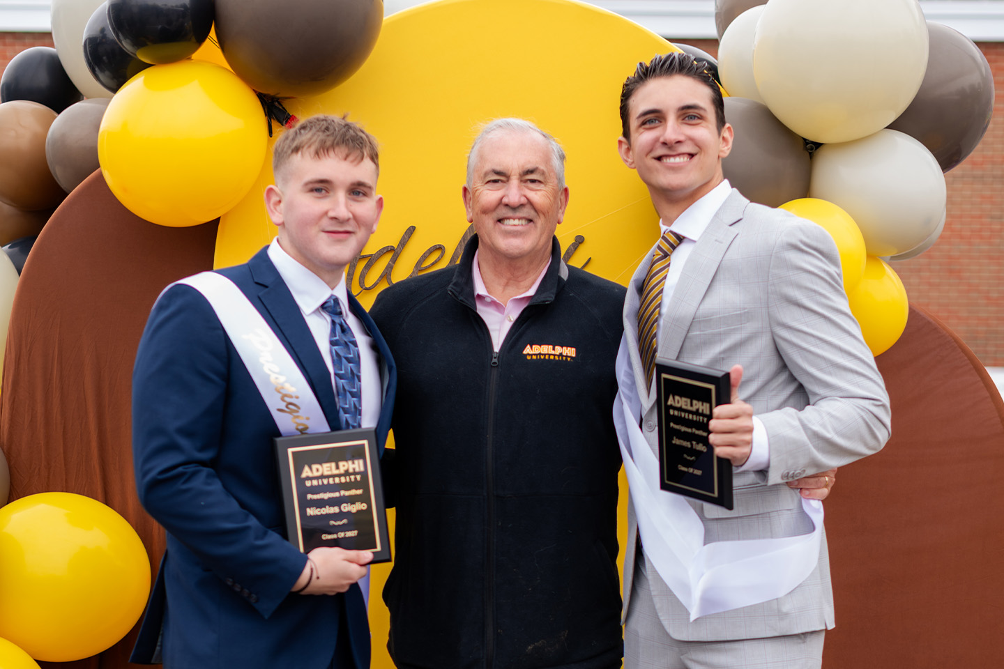 Two young students hold plaques as they stand to either side of Adelphi University President Michael Balboni '81, JD. All three smile proudly.