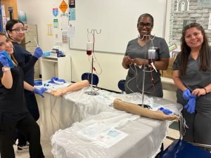 Four students in gray and black scrubs smile at the camera while practicing phlebotomy skills on medical training arm models with IV equipment in a classroom.