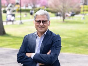 A man wearing a suit and glasses, Adelphi's first Fulbright-Scholar-in Residence, poses in front of the beautiful Adelphi campus in spring time.