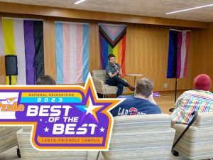 The Campus Pride Best of the Best Badge is displayed over a photograph of students in the Multicultural Center. The badge, which includes a large star as well as four smaller ones, also reads "National Recognition 2023" and "LGBTQ-friendly institution." In the photograph, a student holds a microphone and addresses a group of students, four of whom are seen. Four colorful LGBTQ and trans flags are on the wall behind the speaker.