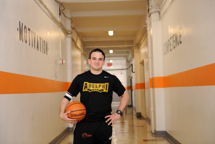 An Adelphi alumni standing confidently in a hallway of a school holding a basketball.