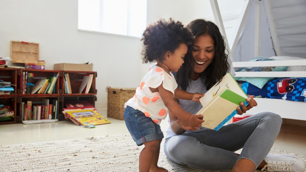Parent smiling with their young child on the floor reading a book