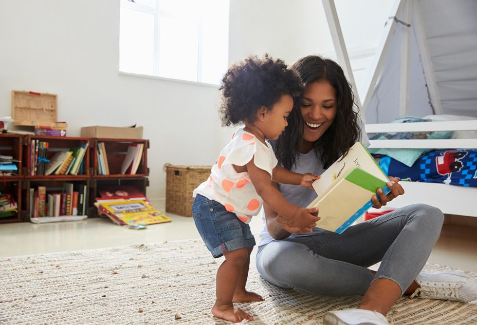 Parent smiling with their young child on the floor reading a book