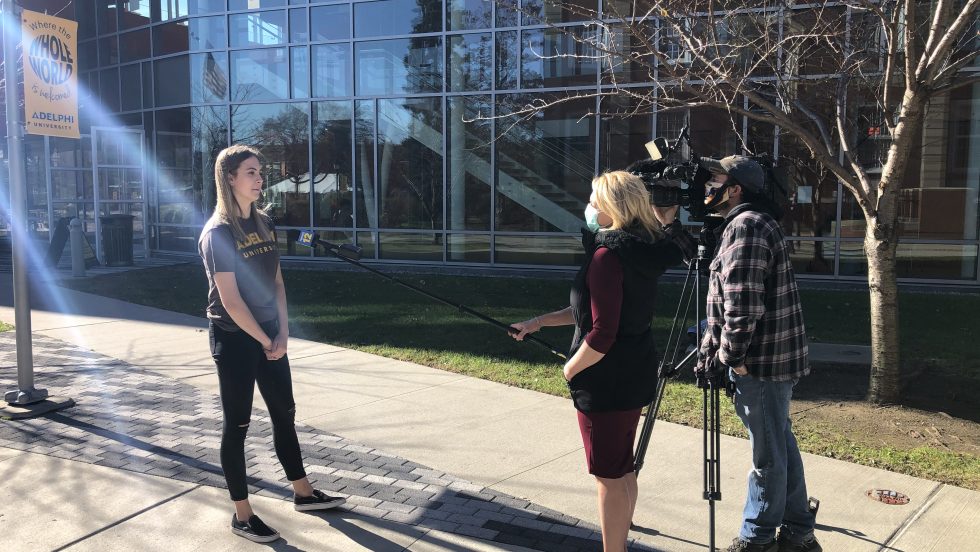 An Adelphi student getting interviewed by news media while social distancing; The microphone is on a pole and the newscaster and camera operator are standing away from the student.