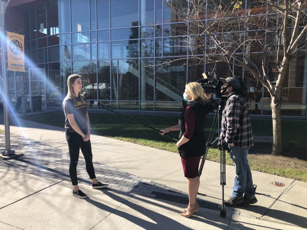 An Adelphi student getting interviewed by news media while social distancing; The microphone is on a pole and the newscaster and camera operator are standing away from the student.