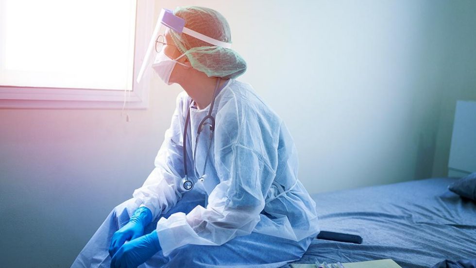 A nurse in PPE sitting on a bed and looking out a window.