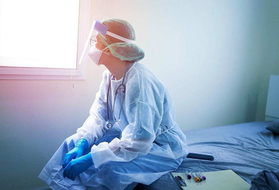 A nurse in PPE sitting on a bed and looking out a window.