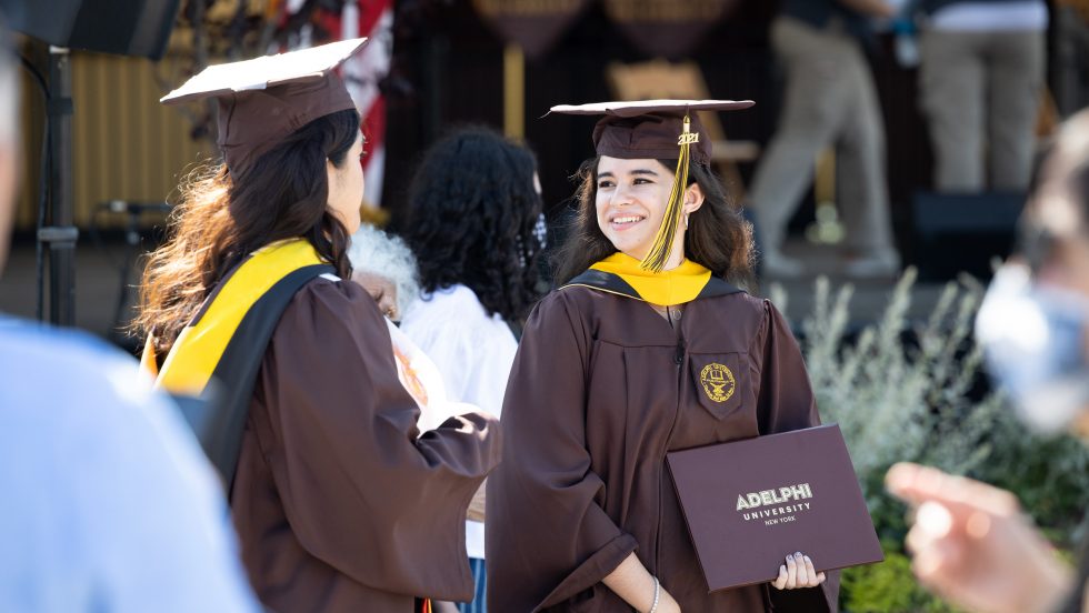 Two Adelphi graduates holding diplomas and wearing regalia.