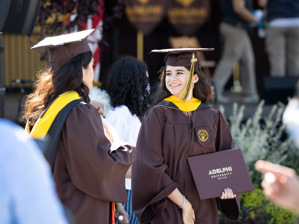 Two Adelphi graduates holding diplomas and wearing regalia.