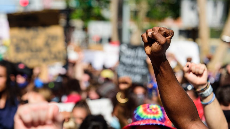 A photo of a racial equality protest focused on the upraised fists. Signs and people are blurred in the background.