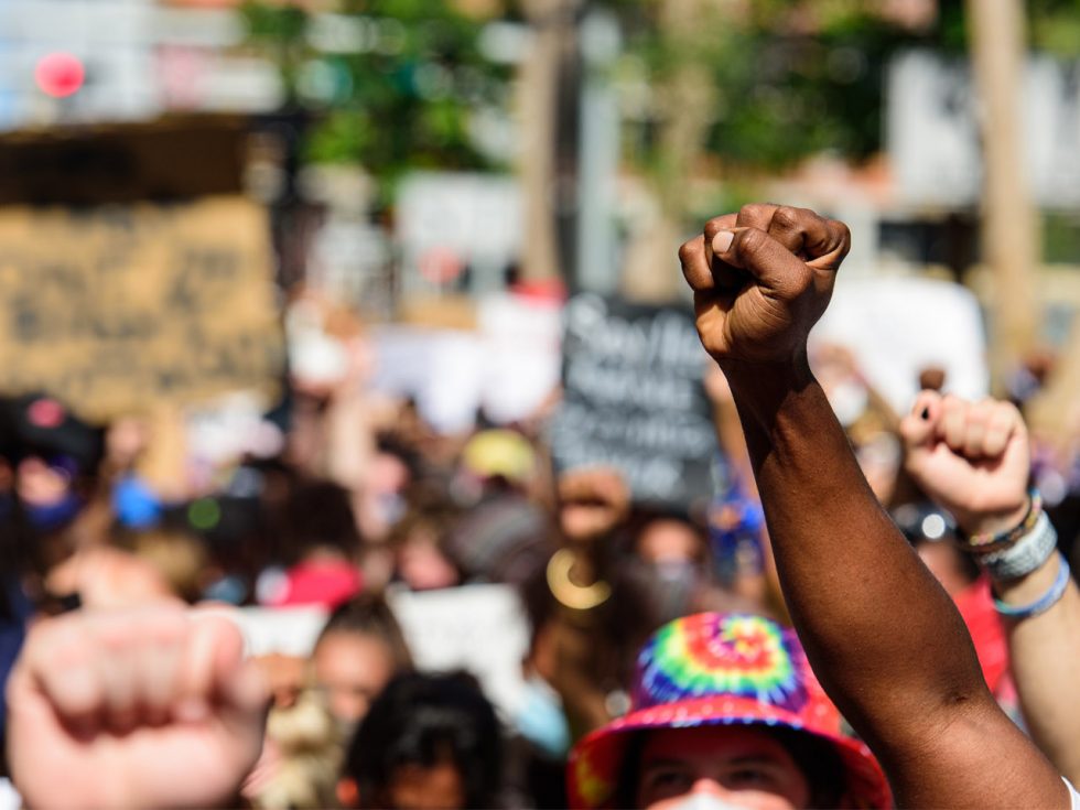 A photo of a racial equality protest focused on the upraised fists. Signs and people are blurred in the background.