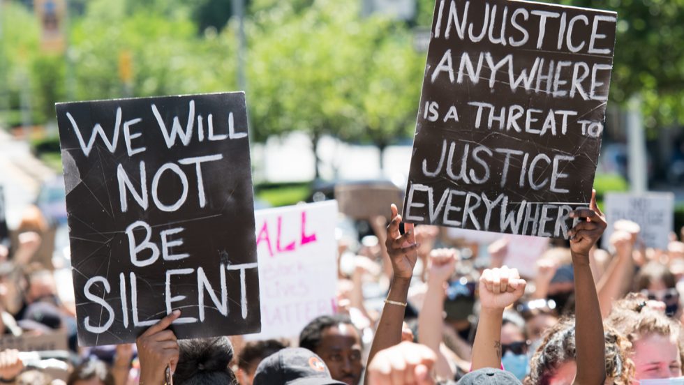 A crowd during a peaceful protest holding signs that say "we will not be silent" and "injustice anywhere is a threat to justice everywhere".