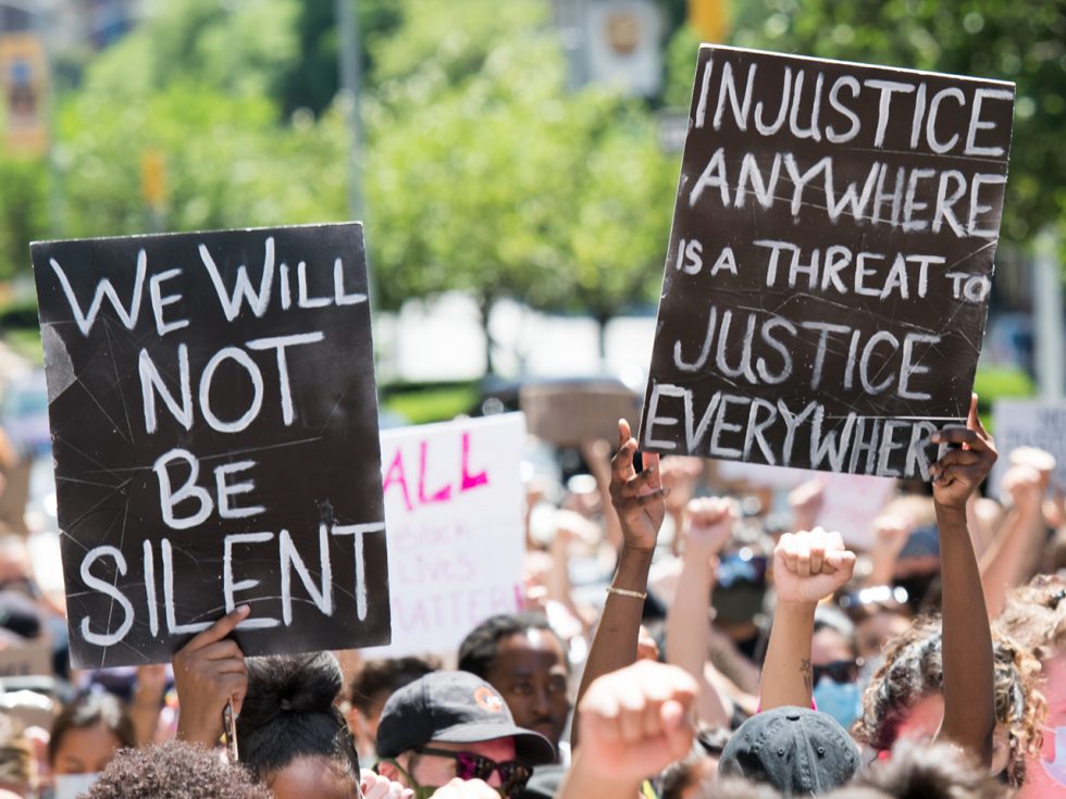 A crowd during a peaceful protest holding signs that say "we will not be silent" and "injustice anywhere is a threat to justice everywhere".