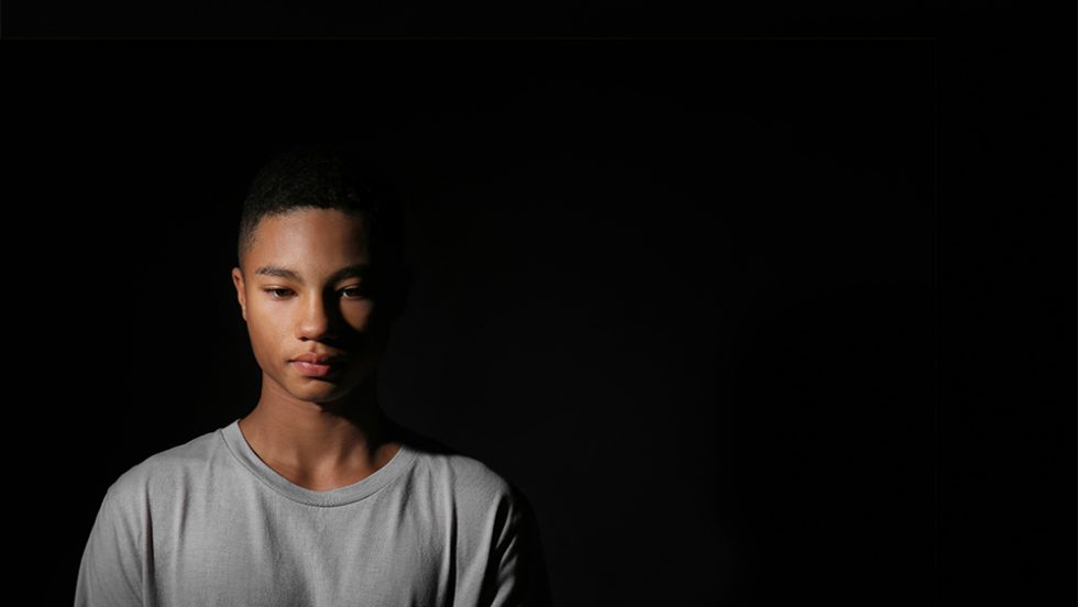 A photo of a young Black man with a somber expression, standing on a stark darkened background.