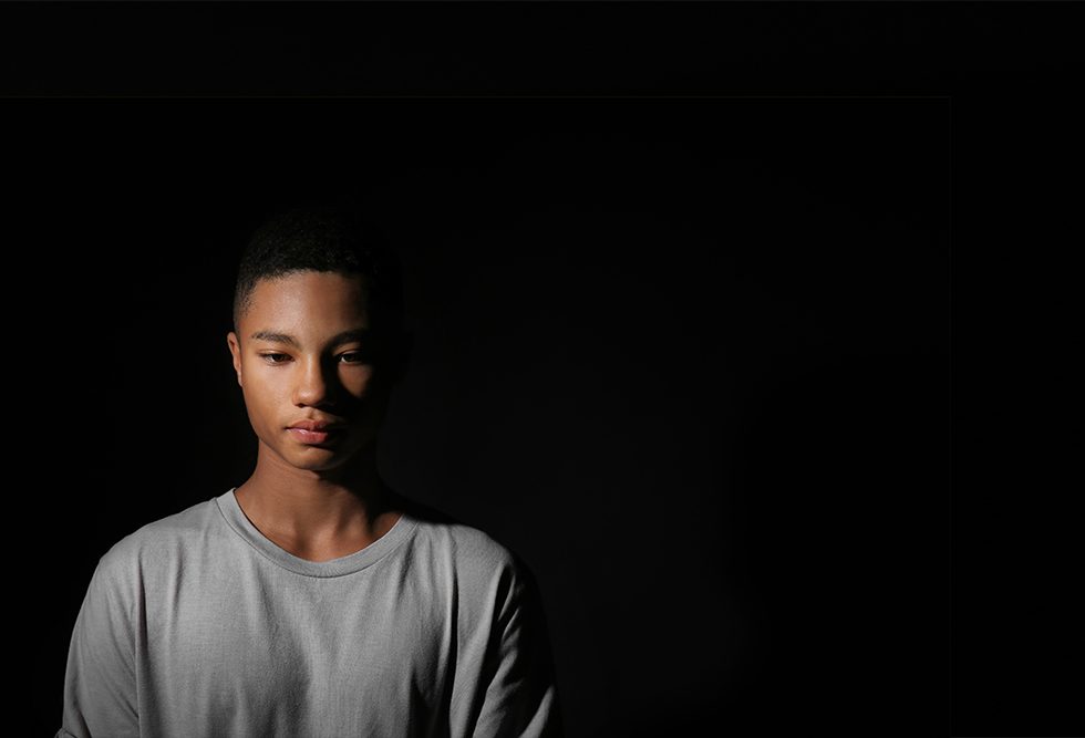 A photo of a young Black man with a somber expression, standing on a stark darkened background.
